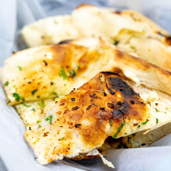 Close-up of golden-brown Indian naan bread, garnished with green herbs, served in a white paper-lined basket.