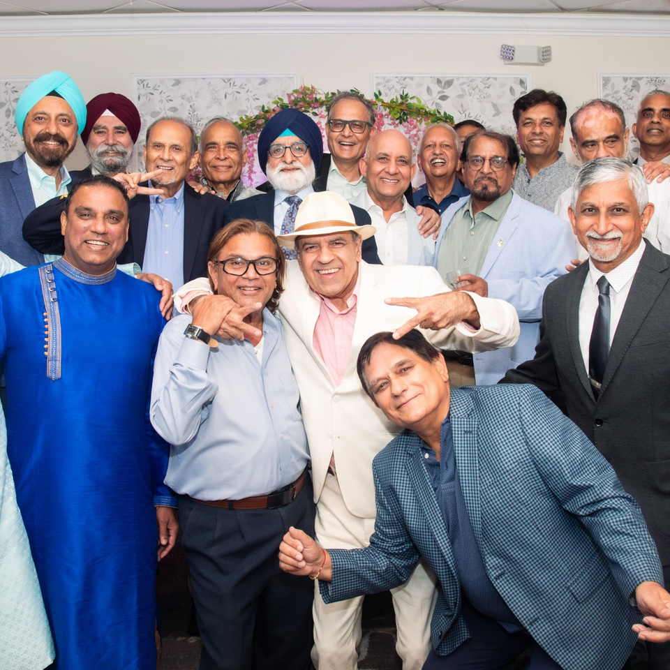 Large group of smiling South Asian men posing together indoors, including two men wearing turbans and one wearing a white fedora.