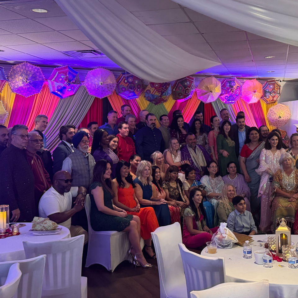 Large group posing at a restaurant celebration beneath colorful draped fabrics, decorative umbrellas, and purple event lighting.